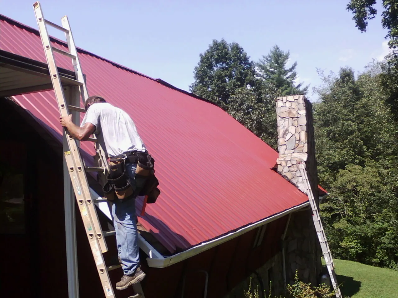 Roofer on a ladder beside a red metal roof and stone chimney — Soffit & Fascia Repair by Roofing Brothers in Biscoe